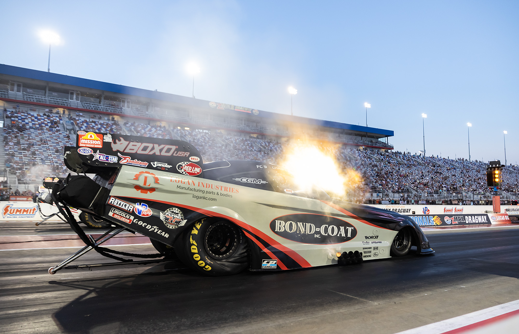 Chad Green's Bond-Coat Funny Car launches with header flames in front of a full grandstand at zMAX Dragway