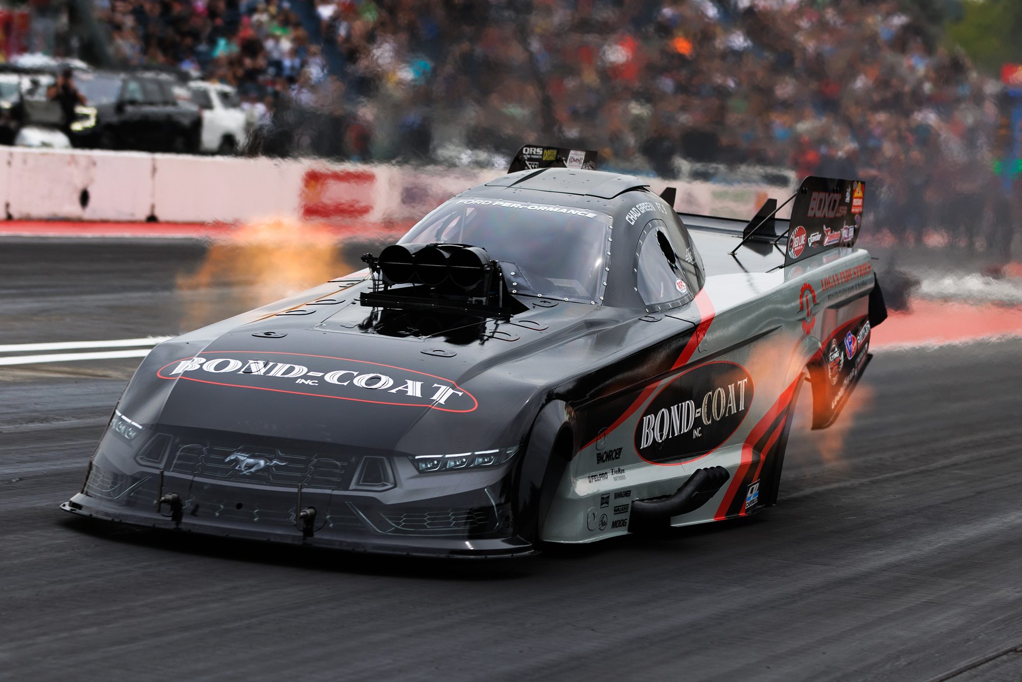 The Bond-Coat Mustang Funny Car at speed, header flames lit, in front of a full grandstand